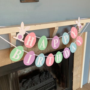 May include: A pastel colored Happy Birthday banner with white letters on each egg-shaped piece. The banner is strung with white string and has two white bunny cutouts.