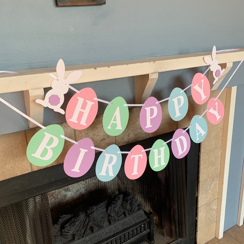 May include: A pastel colored Happy Birthday banner with white letters on each egg-shaped piece. The banner is strung with white string and has two white bunny cutouts.