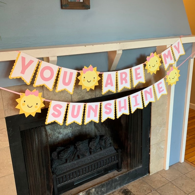 May include: A pink and yellow banner with the text "You are my sunshine" and sun illustrations. The banner is hanging above a fireplace.