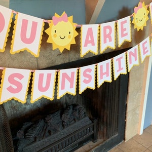 May include: A yellow and pink banner with the words "You Are Sunshine" in pink letters. The banner is decorated with yellow suns with pink bows and smiling faces.