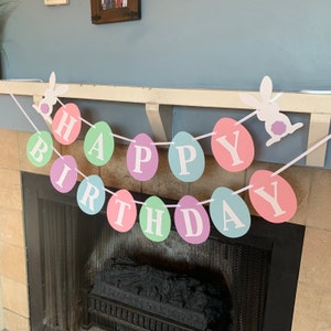 May include: A pastel colored Happy Birthday banner with bunny rabbit cutouts. The banner is made of paper and is strung together with white ribbon. The banner is hanging above a fireplace.