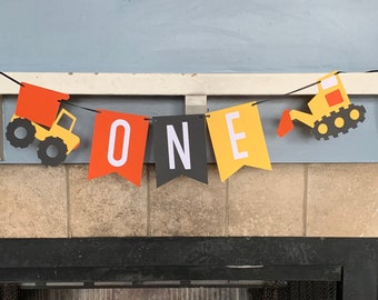 May include: A yellow, orange, and black construction vehicle themed banner with the word "ONE" in black letters. The banner is strung with string and has two construction vehicles on either end.