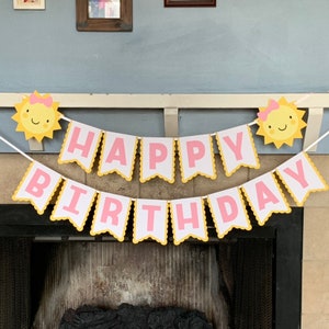 May include: A pink and yellow happy birthday banner with a sun design on each end. The banner is strung up over a fireplace.