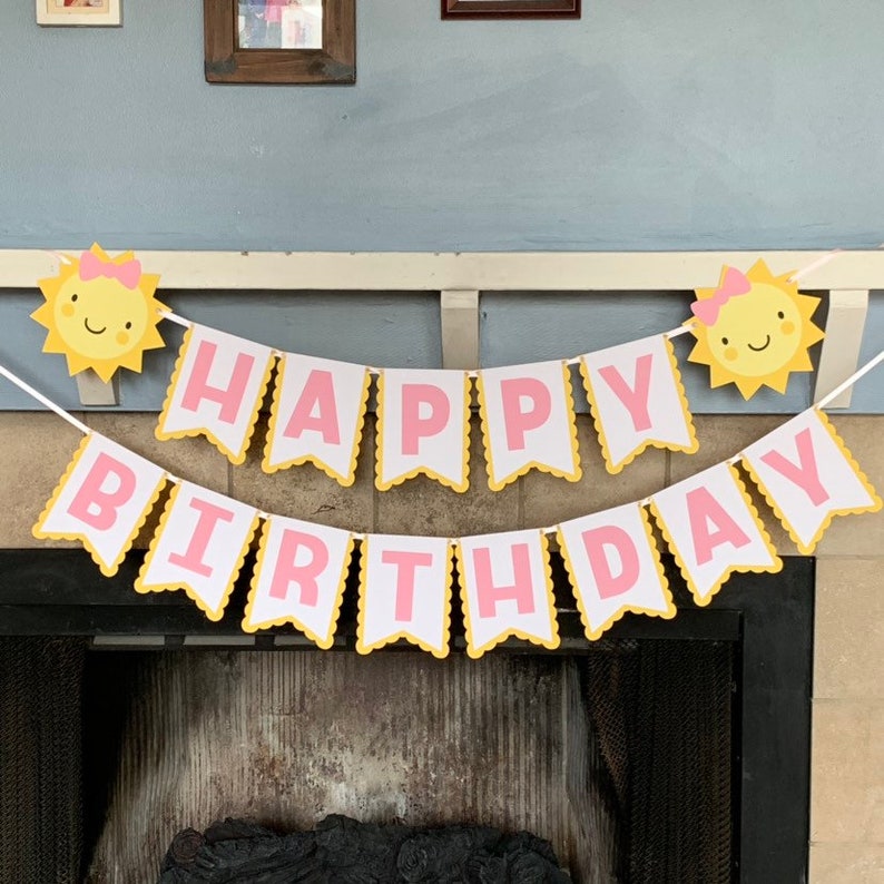 May include: A pink and yellow happy birthday banner with a sun design on each end. The banner is strung up over a fireplace.