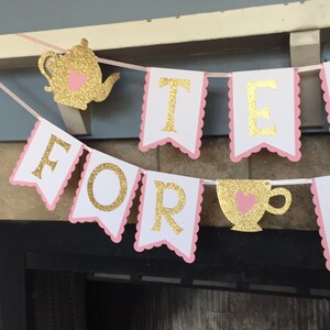 May include: A pink and gold banner with the words "Tea For" and a gold teacup with a pink heart. The banner is decorated with scalloped edges.