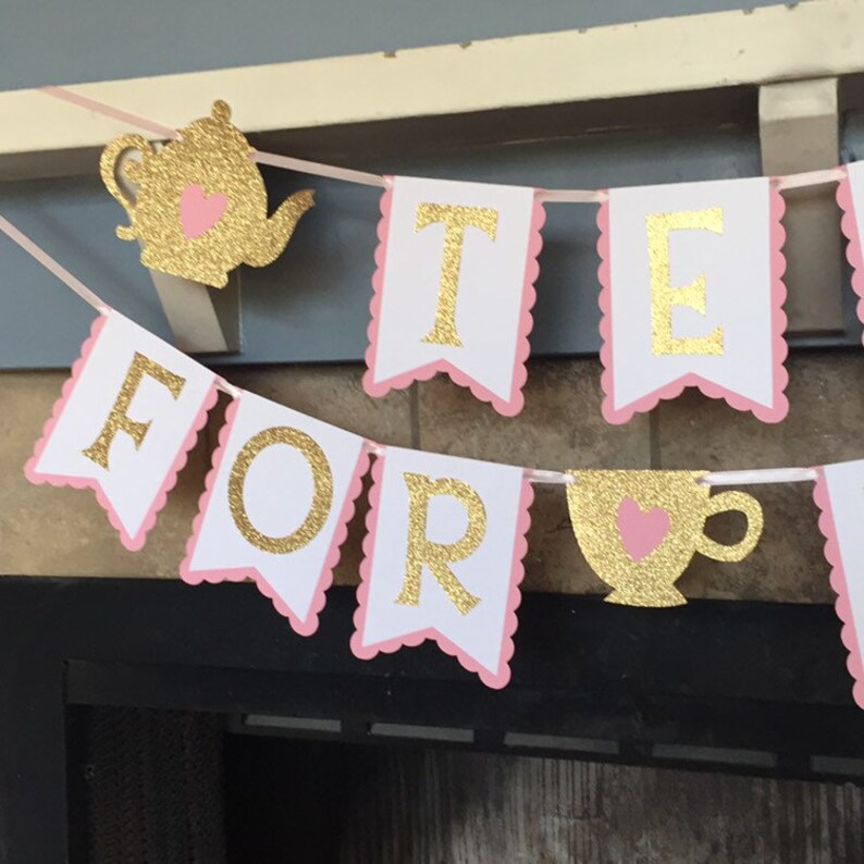 May include: A pink and gold banner with the words "Tea For" and a gold teacup with a pink heart. The banner is decorated with scalloped edges.