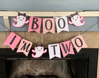 May include: A pink and white banner with the words "BOO I'M TWO" in black glitter lettering. The banner is decorated with ghost-shaped cutouts with black bows. The banner is strung across a fireplace.