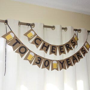 May include: A brown paper banner with black lettering that reads "Cheers to 30 Years". The banner is decorated with small images of beer mugs.