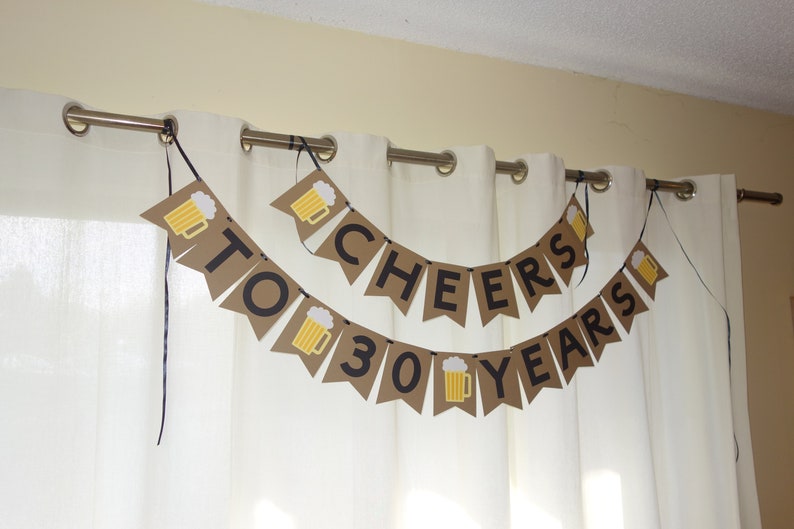 May include: A brown paper banner with black lettering that reads "Cheers to 30 Years". The banner is decorated with small images of beer mugs.