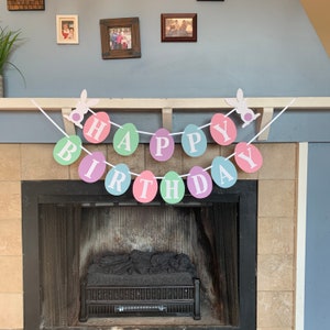 May include: A pastel-colored Happy Birthday banner with bunny rabbit cutouts and white letters on each circle. The banner is hung above a fireplace.