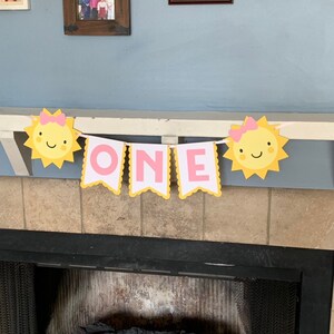 May include: A yellow and pink banner with the word "ONE" in pink letters and two yellow suns with pink bows. The banner is hanging from a white mantle.