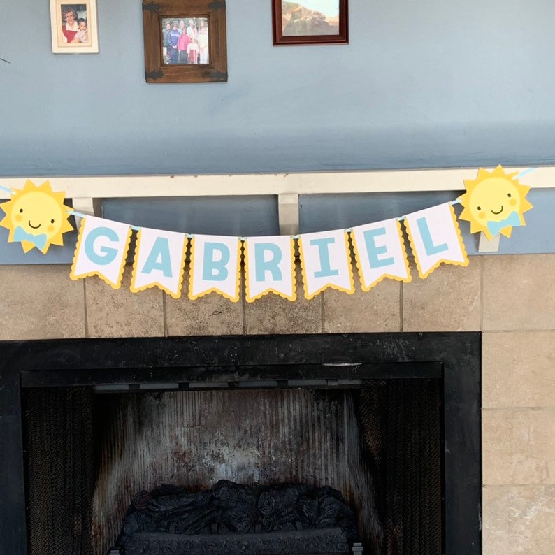 May include: A banner with the name "GABRIEL" in light blue letters on white pennants, trimmed with yellow scalloped edges. The banner is decorated with smiling sun cutouts on each end, creating a cheerful, celebratory atmosphere.