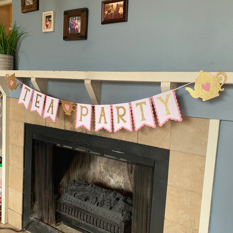 May include: A pink and gold glitter tea party banner with the words "TEA PARTY" in gold letters. The banner is strung up above a fireplace.