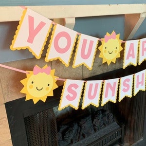 May include: A yellow and pink banner with the words "You Are Sunshine" in pink letters. The banner has a scalloped edge and is decorated with yellow suns with pink bows and smiling faces.