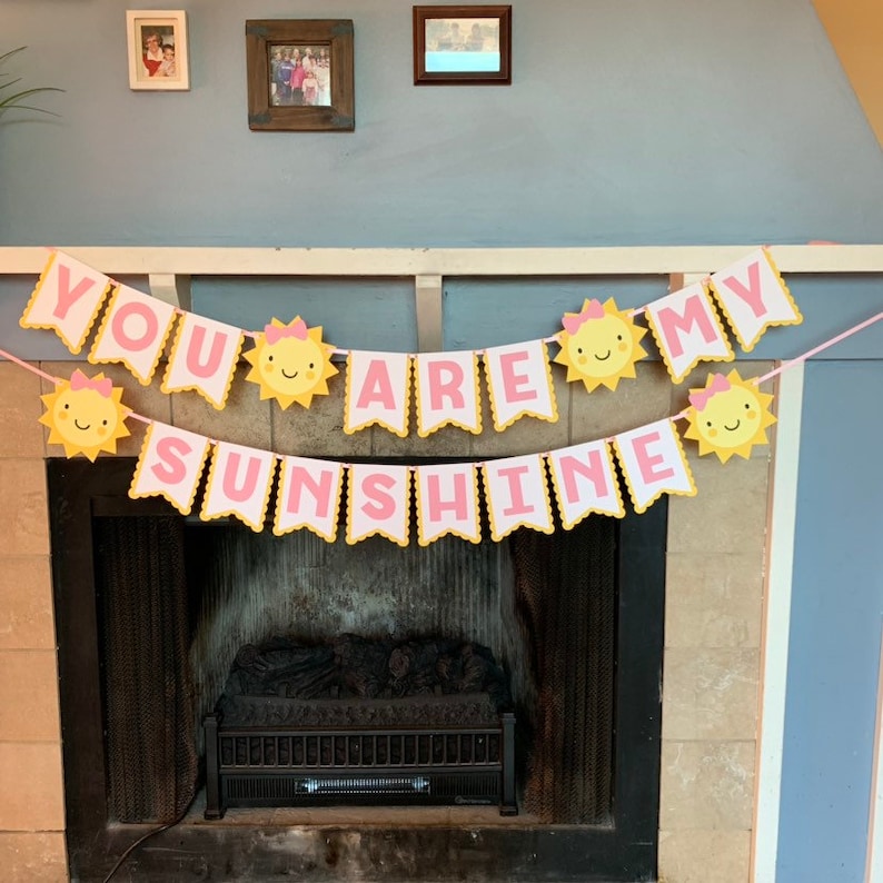 May include: A pink and yellow banner with the words "You Are My Sunshine" in white letters. The banner is decorated with yellow suns with pink bows and smiling faces.