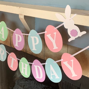 May include: A pastel colored Happy Birthday banner made of paper eggs with a white bunny cutout hanging from the top.