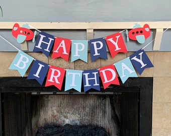 May include: A red, white, and blue banner with the words "Happy Birthday" and airplane cutouts. The banner is hanging above a fireplace.
