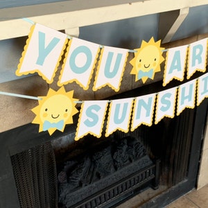 May include: A banner with the words "YOU ARE SUNSHINE" in light blue letters on white pennants with yellow scalloped edges. The banner includes smiling sun illustrations with blue bow ties. The banner is strung across a fireplace.