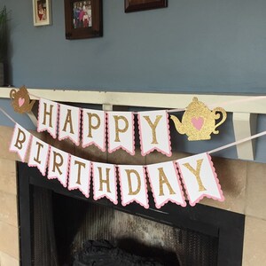 May include: A white and pink banner with gold glitter letters that reads "Happy Birthday". The banner is decorated with two gold glitter teapots with pink hearts.
