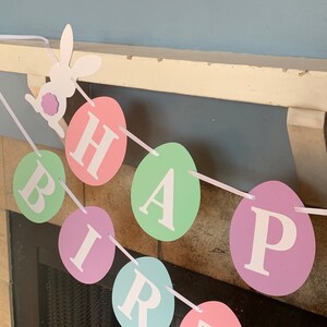 May include: A pastel-colored Easter banner with the word "HAPPY BIRTHDAY" spelled out on individual egg-shaped paper cutouts. A white paper bunny cutout with a purple flower is at the top of the banner.