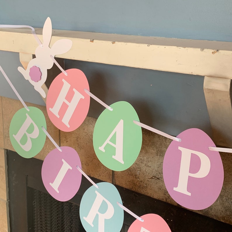 May include: A pastel-colored Easter banner with the word "HAPPY BIRTHDAY" spelled out on individual egg-shaped paper cutouts. A white paper bunny cutout with a purple flower is at the top of the banner.