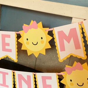 May include: A yellow and pink banner with the word "Sunshine" spelled out in pink letters. Each letter is surrounded by a yellow scalloped border. There are two yellow suns with pink bows and smiling faces on the banner.