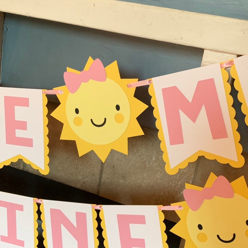 May include: A yellow and pink banner with the word "Sunshine" spelled out in pink letters. Each letter is surrounded by a yellow scalloped border. There are two yellow suns with pink bows and smiling faces on the banner.