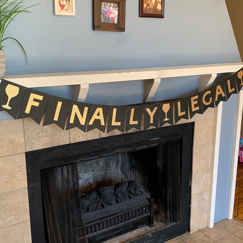 May include: Black and gold "Finally Legal" banner with wine glass icons. The banner is hanging above a fireplace.