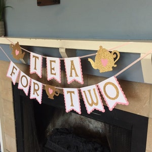 May include: A pink and gold glitter banner with the words "Tea for Two" in white letters. The banner is decorated with teapots and teacups.
