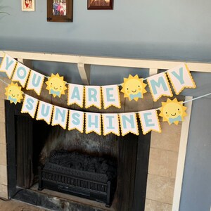 May include: A banner with the text "YOU ARE MY SUNSHINE" in light blue letters. The banner has a scalloped edge and is decorated with yellow suns with smiling faces. The banner is hanging above a fireplace.