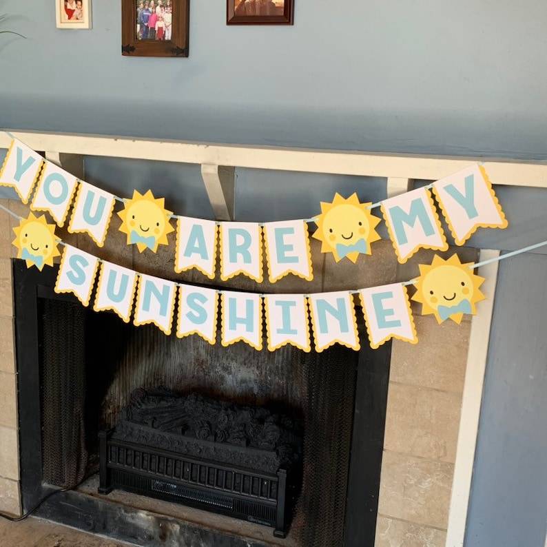 May include: A banner with the text "YOU ARE MY SUNSHINE" in light blue letters. The banner has a scalloped edge and is decorated with yellow suns with smiling faces. The banner is hanging above a fireplace.