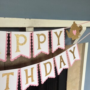 May include: A pink and white banner with gold glitter letters that reads "Happy Birthday". A gold glitter teapot with a pink heart is hanging from the banner.