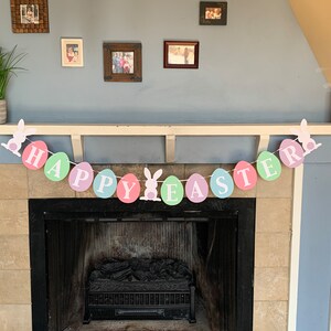 May include: A pastel colored Happy Easter banner with bunny rabbit cutouts hangs above a fireplace. The banner is made of paper circles with the words "Happy Easter" written on them.