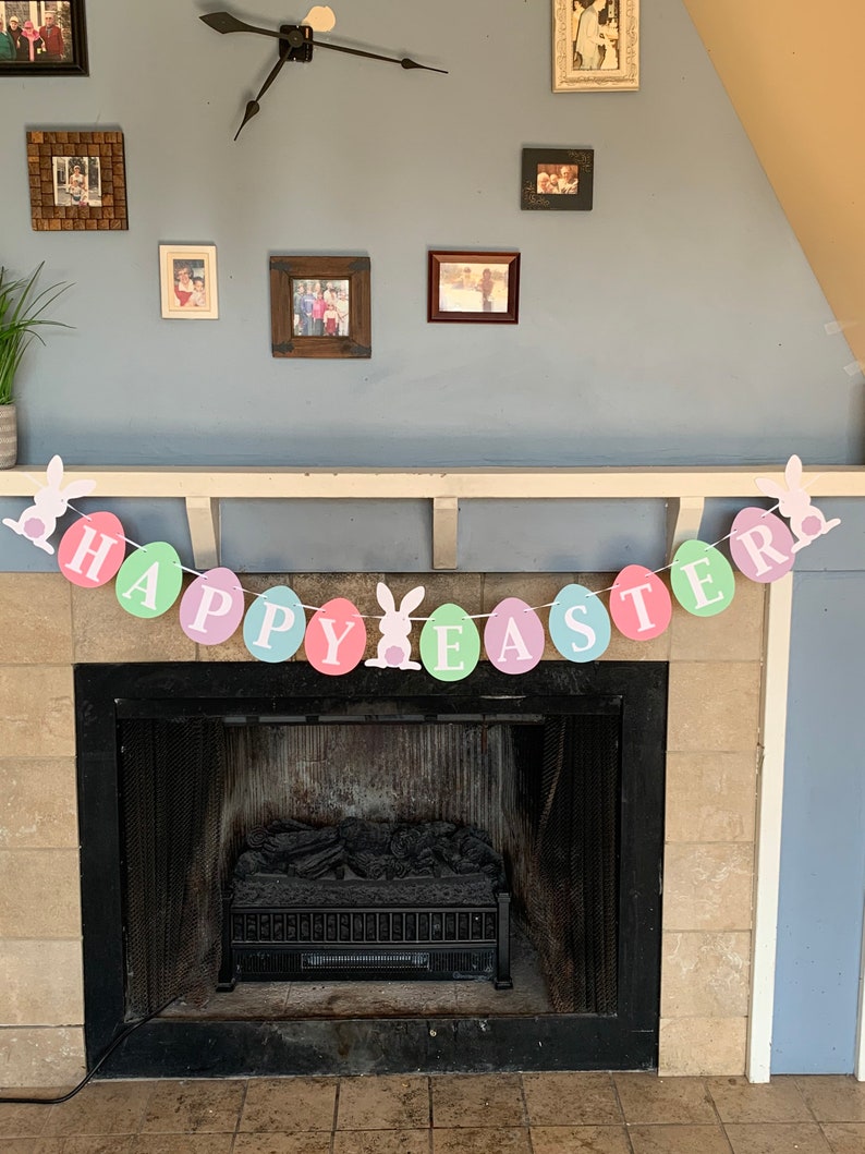 May include: A pastel colored Happy Easter banner with bunny rabbit cutouts hangs above a fireplace. The banner is made of paper circles with the words "Happy Easter" written on them.