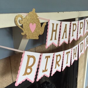May include: A pink and gold banner with the words "Happy Birthday" in gold glitter letters. The banner is decorated with a gold glitter teapot with a pink heart.