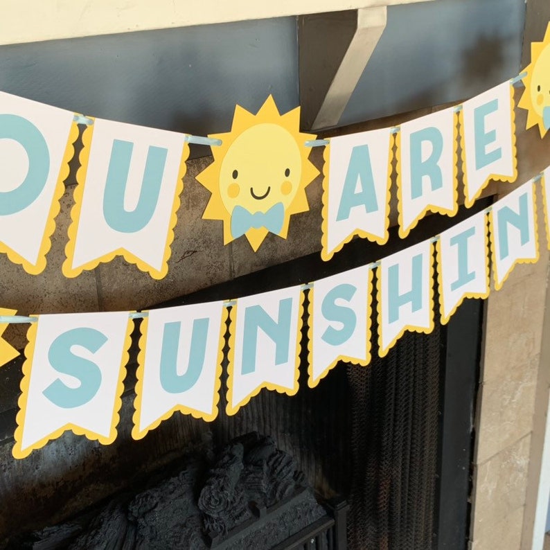 May include: A banner with the text "YOU ARE SUNSHINE" in light blue letters on white pennants with yellow scalloped edges. The banner includes a yellow sun with a blue bow tie.