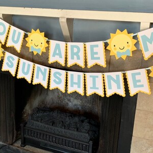 May include: A banner with the words "YOU ARE SUNSHINE" in light blue letters on white pennants. The pennants have a yellow scalloped border and are strung together. Yellow sun illustrations with blue bow ties are also on the banner.