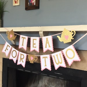 May include: A pink and gold banner with the words "Tea for Two" in gold glitter letters. The banner has pink scalloped edges and features a teapot and teacup with hearts.