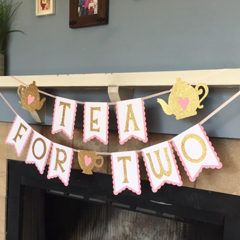 May include: A pink and gold banner with the words "Tea for Two" in gold glitter letters. The banner has pink scalloped edges and features a teapot and teacup with hearts.