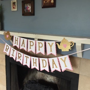 May include: A pink and white banner with gold glitter letters that reads "Happy Birthday". The banner is decorated with two gold glitter teapots with pink hearts.