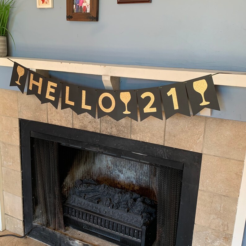 May include: Black and gold banner with the text "HELLO 21" and wine glass icons. The banner is hanging above a fireplace.