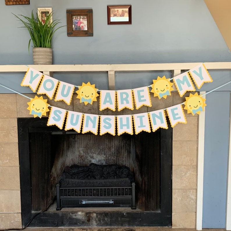May include: A banner with the text "YOU ARE MY SUNSHINE" in light blue letters. The banner features yellow sun illustrations with smiling faces. The banner is strung across a fireplace, creating a cheerful and decorative display.