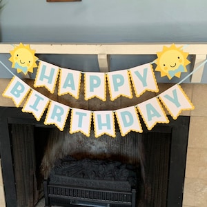 May include: A festive "Happy Birthday" banner with a cheerful sun design. The banner features white pennants with blue lettering and a yellow scalloped border. Two smiling sun cutouts flank the banner, creating a bright and celebratory decoration.