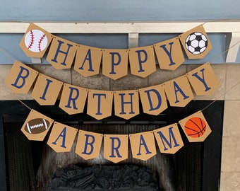 May include: A brown banner with the words "Happy Birthday Abram" in blue letters. The banner is decorated with various sports balls, including a baseball, soccer ball, football, and basketball.