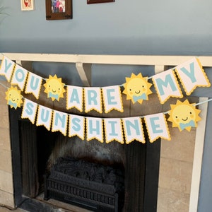 May include: A two-tiered banner with the words "YOU ARE MY SUNSHINE" in blue letters on white pennants. The banner is decorated with yellow sun-shaped cutouts with smiling faces and blue bow ties. The banner is hanging above a fireplace.
