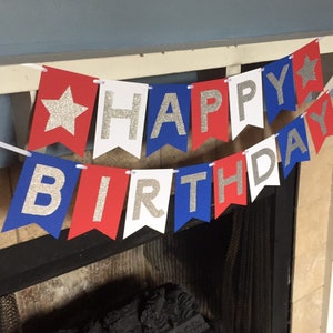 Puede incluir: Bandera roja, blanca y azul con letras de purpurina plateada que deletrean "Happy Birthday". La bandera tiene estrellas en los extremos.