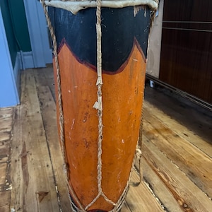 May include: A large, wooden drum with a natural leather head. The drum is painted with black and orange stripes and has a rope tied around it.