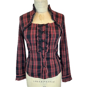 May include: A long-sleeved button-down shirt featuring a red, black, and white plaid pattern. The shirt includes a ruffled collar, a vertical ruffled placket down the center front, and ruffled cuffs. The garment is displayed on a mannequin.