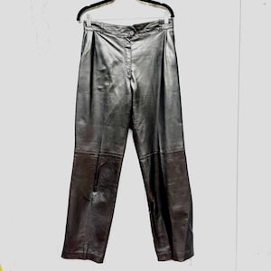 May include: Black leather pants with a button closure and pleats. The pants are hanging on a black hanger. The leather has a slight sheen.
