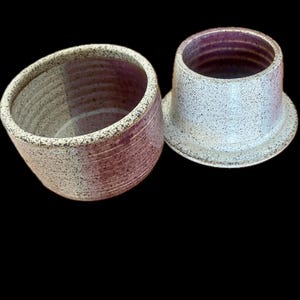 May include: A two-piece ceramic butter keeper featuring a speckled off-white exterior with a purple-toned interior. The set includes a deep bowl-shaped base with horizontal ridges and a matching lid with a wide rim, both finished in a speckled glaze.